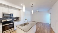 Located in Austin, TX, this thoughtfully designed 2-bedroom, 2-bathroom residence offers a clean, modern aesthetic across a well-organized single-floor layout. The open-concept kitchen and living area serves as the heart of the home, anchored by a sleek center island with a stainless steel sink and quartz-style countertops. The kitchen is fully equipped with stainless steel appliances—including a refrigerator, electric range, and microwave—complemented by crisp white cabinetry extending to the ceiling and a striking dark tile backsplash. Pendant lighting above the island adds a contemporary touch. The living area extends naturally from the kitchen, bathed in natural light from large windows and finished with warm-toned wood flooring that runs throughout the entire space. A ceiling fan provides added comfort. Both bedrooms are bright and well-proportioned, with natural light filtering through slatted blinds. The primary bedroom includes access to a dedicated walk-in closet with built-in shelving. The bathrooms feature white vanities, mirrors with overhead lighting, and matching wood-look flooring. Clean white walls, recessed lighting, and a consistent modern palette give the entire residence a fresh, move-in-ready feel.