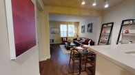 This single-floor residence is designed with an open-concept layout that seamlessly connects the dining and living areas, creating a cohesive and functional living space. Rich, dark hardwood flooring runs throughout, providing a warm and grounded foundation that complements the neutral wall tones of beige and soft gray.

The dining area, positioned near the kitchen peninsula, features a glass-top table with stylish wooden chairs, making it an intimate space for everyday meals. A lean, open shelving ladder unit nearby adds a decorative touch while offering practical storage.

Beyond an arched entryway, the living room opens up spaciously, anchored by a dark leather sofa accented with warm red and earthy-toned throw pillows. A glass coffee table sits atop a layered area rug, adding texture and definition to the seating zone. Two windows dressed with white blinds allow natural light to filter in, brightening the space.

Wall art in bold reds and cool blues adds personality and visual interest throughout. Recessed lighting provides consistent illumination. A secondary seating area with a red accent chair near the windows adds depth and versatility to the overall layout.