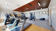 This contemporary fitness center is thoughtfully designed to support a wide range of workout routines. The space is defined by its standout architectural feature: a slatted warm-wood ceiling that spans the central zone of the gym, adding visual depth and a refined aesthetic contrast to the crisp white walls. Linear pendant lighting integrated into the ceiling structure ensures the space is well-lit throughout.

The cardio area lines one side of the facility, featuring a row of treadmills and elliptical machines positioned alongside large windows that flood the zone with natural light and offer views to the outdoors. The light hardwood flooring in this section transitions to a patterned carpet in the strength training area, visually delineating the two zones.

The strength training section is comprehensively equipped with cable machines, a smith machine with a weight bench, a dumbbell rack with a full range of free weights, and additional benches for varied exercises. A dedicated open-floor area provides space for functional training, including a tire flip station.

Framed artwork decorates the walls, contributing to a polished, welcoming atmosphere. The facility also includes an accessible water fountain and restrooms conveniently located adjacent to the workout floor, completing a fully functional and well-appointed fitness environment.