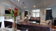 This well-designed single-floor space makes a strong impression with its open and airy layout. The main living area features rich hardwood flooring that transitions to light tile, creating a welcoming contrast underfoot. Vaulted ceilings add a generous sense of height and volume to the room, while recessed lighting and a ceiling fan provide both ambiance and comfort. Large windows and glass-paned French doors allow natural light to pour in, offering views of the lush greenery outside and brightening the entire space. The living area is elegantly furnished with deep-toned upholstered sofas accented by patterned throw pillows, complemented by ornate accent chairs and a sleek glass-top coffee table. Decorative wall art panels add a sophisticated, textured element to the soft blue-grey walls. A striking floral arrangement in a glass vase serves as a bold focal point, lending a touch of organic color and warmth. A separate seating nook with a classic floor lamp creates an intimate corner for reading or conversation. The overall ambiance is refined yet inviting, blending contemporary style with comfortable, livable design throughout.