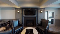 This well-appointed single-floor amenity lounge is designed to offer a refined social and recreational environment. The space is anchored by a sophisticated lounge area centered around a tiled fireplace framed by built-in paneling and flanked by a wall-mounted flat-screen television and elegant sconce lighting. A pair of deep leather armchairs and a round coffee table complete the intimate seating arrangement, creating a welcoming gathering spot.

Adjacent to the lounge area, a billiards table serves as a central feature, providing a focal point for casual recreation. Decorative columns define distinct zones throughout the open floor plan without compromising the sense of flow and connectivity between areas.

A high-top dining area positioned near large windows with horizontal blinds offers an abundance of natural light, with views of the exterior surroundings. The bar-height table and mix of leather and upholstered stools give this corner a relaxed, social atmosphere. A banquette seating area with tufted upholstery adds an additional intimate dining or gathering option.

Throughout the space, warm-toned hardwood flooring ties the various zones together cohesively. Tray ceilings with recessed lighting add architectural depth, while the cool grey and blue color palette lends the overall environment a contemporary, polished character. A glass-paneled entry door allows natural light to filter into the space.