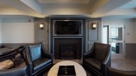 This well-appointed single-floor amenity lounge is designed to offer a refined social and recreational environment. The space is anchored by a sophisticated lounge area centered around a tiled fireplace framed by built-in paneling and flanked by a wall-mounted flat-screen television and elegant sconce lighting. A pair of deep leather armchairs and a round coffee table complete the intimate seating arrangement, creating a welcoming gathering spot.

Adjacent to the lounge area, a billiards table serves as a central feature, providing a focal point for casual recreation. Decorative columns define distinct zones throughout the open floor plan without compromising the sense of flow and connectivity between areas.

A high-top dining area positioned near large windows with horizontal blinds offers an abundance of natural light, with views of the exterior surroundings. The bar-height table and mix of leather and upholstered stools give this corner a relaxed, social atmosphere. A banquette seating area with tufted upholstery adds an additional intimate dining or gathering option.

Throughout the space, warm-toned hardwood flooring ties the various zones together cohesively. Tray ceilings with recessed lighting add architectural depth, while the cool grey and blue color palette lends the overall environment a contemporary, polished character. A glass-paneled entry door allows natural light to filter into the space.