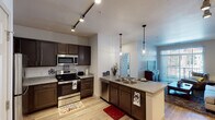 This single-floor residence offers a thoughtfully designed layout that connects the kitchen, living area, bedroom, and bathroom in a functional flow. The kitchen is a standout feature, outfitted with rich dark-brown cabinetry, granite-style countertops, and a full suite of stainless steel appliances including a refrigerator, electric range, microwave, and dishwasher. Light wood-style flooring runs throughout, and track lighting overhead ensures bright, even illumination. A peninsula with a double sink and pendant lighting above adds both workspace and visual interest, opening naturally toward the dining and living areas. The living room is bright and comfortable, featuring a sliding glass door with vertical blinds that provides access to a private balcony and invites natural light into the space. The bedroom offers plush carpeting, a ceiling fan, and dual windows that bring in additional daylight and exterior views. The bathroom includes a combined shower and tub enclosure with white tile surround, offering a clean and practical finish. Overall, the space presents a modern, cohesive aesthetic suited for comfortable everyday living.