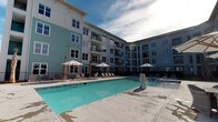 Located at 2155 Morris Basket Blvd in North Charleston, South Carolina, this contemporary residential community showcases a striking multi-story building with a well-designed exterior finished in a combination of soft teal, gray, and white tones. The architecture features clean lines, large windows, and private balconies that overlook the central courtyard area below.

At the heart of the community is a generously sized resort-style swimming pool, serving as a focal point for outdoor living. The pool deck is finished in light-toned concrete and is surrounded by an array of lounge chairs and market umbrellas, creating an inviting atmosphere for relaxation. Tropical-style palm trees and landscaping accent the poolside environment, contributing to the warm, coastal ambiance characteristic of the South Carolina Lowcountry.

The building's design wraps around the pool courtyard in an L-shaped configuration, providing residents with direct sightlines to the amenity space from their balconies. The overall aesthetic is modern and resort-inspired, balancing comfort and contemporary design in a thoughtfully planned outdoor setting.