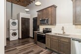 This single-floor loft-style unit showcases a compelling blend of industrial character and contemporary design. Soaring ceilings with exposed wooden beams and open rafters lend a raw, architectural warmth throughout the living areas, while large windows flood the space with generous natural light, casting bright patterns across the floors.

The kitchen is a standout feature, outfitted with rich, dark-stained shaker-style cabinetry, granite countertops, and a classic white subway tile backsplash. Stainless steel appliances — including a range, microwave, dishwasher, and refrigerator — complement the space's modern sensibility, while pendant lights hang from the exposed beam ceiling above, adding an industrial-chic touch. Wood-look plank flooring runs throughout the kitchen and main living areas, enhancing the loft aesthetic.

The bedrooms offer a quieter atmosphere, carpeted for comfort, with large windows that bring in abundant daylight and ceiling fans overhead. The exposed brick and open rafter details visible in these rooms reinforce the building's industrial heritage.

Both bathrooms are cleanly finished with walk-in showers featuring white surrounds, granite-topped vanities with dark wood cabinetry, and wood-look flooring. One bathroom also includes built-in open shelving for added storage. The overall ambiance is modern and refined, with nods to the building's historic industrial roots at every turn.