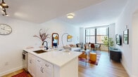 Located on the 7th floor at 100 Christopher Columbus Dr in Jersey City, NJ, this one-bedroom, one-bathroom residence offers a thoughtfully designed layout across a single floor. Light-toned hardwood flooring flows throughout, complementing the clean white walls and creating a bright, cohesive aesthetic.

The open-concept living and dining area is anchored by oversized, floor-to-ceiling windows that frame sweeping city views and flood the space with natural light. The kitchen features white shaker cabinetry, stainless steel appliances, a subway tile backsplash, and a generous white countertop island with bar seating — ideal for both cooking and entertaining.

The bedroom is a comfortable retreat with large windows overlooking the urban skyline, offering ample space for a queen bed, workspace, and storage. The room is finished with warm, neutral tones and connects to a dedicated closet.

The bathroom is clean and modern, appointed with hexagonal floor tile, a white vanity with black fixtures, a framed mirror, and a tiled shower-tub combination. A hallway provides a functional transition between rooms, completing the efficient and well-organized floor plan. This residence combines contemporary finishes with practical urban living in the heart of downtown Jersey City.