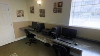 This single-floor business center offers a practical and organized workspace designed to meet everyday computing needs. A long granite-topped counter runs along the walls, providing ample desk space for multiple users simultaneously. The workstations are equipped with desktop computers, keyboards, and monitors, along with a shared laser printer conveniently positioned for easy access. Ergonomic rolling office chairs accompany each station, ensuring comfort during extended use. Natural light filters into the room through a window fitted with horizontal blinds, creating a bright yet adjustable atmosphere. The walls are painted in a warm neutral tone and accented with a chair rail molding, lending a polished, professional feel to the space. Decorative wall art featuring textured square panels adds a touch of style and visual interest throughout the room. A wall-mounted light fixture provides additional ambient lighting. The space is finished with wall-to-wall carpeting, contributing to a quiet and focused environment. Overall, the business center is thoughtfully arranged to provide a productive and welcoming setting for work, research, or communication tasks.