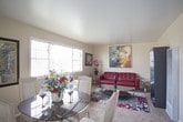 This single-floor residence offers a cohesive and thoughtfully designed living environment across several distinct spaces. The living room serves as the heart of the home, featuring plush wall-to-wall carpeting and a bold red tufted leather sofa paired with a classic white accent chair. A large, vibrant abstract painting anchors the main wall, complemented by additional framed artwork that adds depth and personality to the space. A glass-top coffee table and sleek side tables complete the room, while a wide window fitted with horizontal blinds allows for generous natural light and views of the surrounding area. The kitchen is practical and efficient, featuring dark hardwood-style flooring, white cabinetry, and white appliances including a freestanding electric range. A secondary door in the kitchen provides direct exterior access, and a window above the sink brings in additional natural light. The bedroom is a serene retreat, appointed with soft carpeting, a ceiling fan with a light fixture, and a large upholstered tufted headboard in a metallic silver finish. A coordinating dresser and nightstands in a mirrored silver style add elegance, while a window with blinds allows for controlled natural light. Decorative framed artwork and personal accents throughout the home create a warm, curated ambiance.