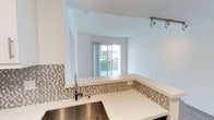 This single-floor space showcases a clean, modern design characterized by bright white walls and an open-concept layout that seamlessly connects the kitchen and living areas. The kitchen is well-appointed with crisp white countertops, a stainless steel sink, and a stylish mosaic tile backsplash featuring neutral tones of grey and beige that adds texture and visual interest. A raised breakfast bar defines the transition between the kitchen and the living space, making it ideal for casual dining or entertaining. Track lighting on the ceiling provides focused illumination throughout the kitchen area. The living space benefits from an abundance of natural light, streaming in through a large sliding glass door that opens to a private outdoor balcony, offering a seamless indoor-outdoor connection. The sliding door frames a pleasant outdoor view, enhancing the airy, open ambiance of the interior. The overall design palette is neutral and contemporary, with consistent finishes throughout that create a cohesive and inviting atmosphere. The thoughtful layout maximizes the use of space while maintaining an uncluttered, functional feel throughout.