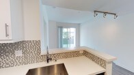 This single-floor space showcases a clean, modern design characterized by bright white walls and an open-concept layout that seamlessly connects the kitchen and living areas. The kitchen is well-appointed with crisp white countertops, a stainless steel sink, and a stylish mosaic tile backsplash featuring neutral tones of grey and beige that adds texture and visual interest. A raised breakfast bar defines the transition between the kitchen and the living space, making it ideal for casual dining or entertaining. Track lighting on the ceiling provides focused illumination throughout the kitchen area. The living space benefits from an abundance of natural light, streaming in through a large sliding glass door that opens to a private outdoor balcony, offering a seamless indoor-outdoor connection. The sliding door frames a pleasant outdoor view, enhancing the airy, open ambiance of the interior. The overall design palette is neutral and contemporary, with consistent finishes throughout that create a cohesive and inviting atmosphere. The thoughtful layout maximizes the use of space while maintaining an uncluttered, functional feel throughout.