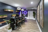 Located at 1701 Forest Ave in Knoxville, Tennessee, this single-floor amenity space offers a thoughtfully designed collection of resident-focused areas that blend modern aesthetics with practical functionality. The centerpiece is a vibrant lounge and bar area, featuring a bold navy hexagonal tile accent wall adorned with rustic wood floating shelves filled with greenery. A sleek dark bar top with industrial-style barstools anchors the space, while eye-catching pendant lighting and a neon ring art installation create a lively, social atmosphere. Light wood-look flooring flows throughout, tying each zone together seamlessly. The fitness center is generously equipped with treadmills, ellipticals, stationary bikes, rowing machines, a punching bag, and cable machines, all set against a dramatic floral mural wall and large windows that bring in natural light and outdoor views. A dedicated package locker room features a striking cityscape mural wallcovering, providing secure and convenient parcel pickup. The business center offers multiple iMac workstations set on wood-and-metal desks beneath a large wall-mounted TV on a reclaimed wood panel, ideal for remote work or study. Additional utility areas and corridors with exposed ductwork and curated wall art complete the industrial-chic character of this well-appointed amenity floor.