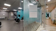 Located at 250 Main St in Hartford, Connecticut, this fitness center offers a well-appointed, single-floor layout designed to support a comprehensive workout experience. The main gym area features durable rubber flooring in a speckled gray finish, providing a safe and functional surface for exercise. The space is equipped with Life Fitness cardio machines, including treadmills, ellipticals, and stationary bikes, as well as a multi-functional cable machine and a full rack of dumbbells for strength training. Teal accent walls and recessed panel ceilings with flush LED lighting give the gym a modern, energetic aesthetic. Glass partitions allow for an open sightline throughout the space, enhancing both safety and the sense of openness. Adjacent to the main gym floor is a dedicated group exercise or yoga studio, separated by floor-to-ceiling glass walls. This room features light wood-look flooring, large wall mirrors, pendant and recessed lighting, and a striking full-wall nature-inspired mural depicting a close-up of a succulent plant, creating a calming and focused atmosphere ideal for stretching, yoga, or fitness classes. Together, the two areas form a cohesive, professionally designed fitness facility suited for a variety of workout routines.