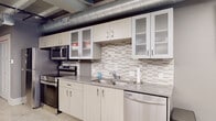 This thoughtfully designed single-floor amenity space blends industrial character with contemporary finishes to create a versatile, welcoming environment. Polished terrazzo flooring runs throughout, reflecting the geometric LED linear lighting suspended from an exposed ceiling that showcases structural beams and large-diameter HVAC ductwork — hallmarks of an industrial-modern aesthetic.

The open-plan layout connects several distinct functional zones. A fully equipped kitchen features stainless steel appliances including a range, over-the-range microwave, dishwasher, and refrigerator, complemented by light wood cabinetry with frosted glass uppers and a textured tile backsplash. Adjacent to the kitchen, a dining area accommodates group seating around a large communal table.

A comfortable lounge area is anchored by a bold geometric-patterned feature wall, plush gray sofas, accent chairs, and a wall-mounted flat-screen television — ideal for relaxed gatherings. A second meeting or dining zone near a window with exterior views offers additional flexible seating and another mounted display screen.

A corridor lined with paneled doors leads through the space, with an electrical panel and private rooms accessible along the way. The restroom is clean and modern, featuring a floating wood vanity, backlit mirror, under-mount sink, and accessibility grab bars. Lush indoor plants add warmth throughout.