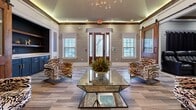 Located at 3085 Commonwealth Dr in Spring Hill, TN, this well-appointed single-floor space offers a thoughtfully designed layout spanning a comfortable footprint. The main living area makes a striking first impression with a tray ceiling accented by warm ambient LED lighting, shiplap-style walls, large windows that invite natural light, and sliding barn doors crafted from rich wood. Bold, patterned accent chairs surround a glass coffee table, while a built-in wet bar with navy cabinetry, open wood shelving, and gold hardware adds a sophisticated entertaining touch. Wood-look tile flooring flows throughout, lending warmth and visual continuity. A dedicated fitness room features mirrored walls, a wall-mounted television, yoga mats, kettlebells, and natural light from a bright window — ideal for workouts or wellness routines. The media lounge showcases grey shiplap walls adorned with classic movie posters, a large wall-mounted screen, plush area rugs, and relaxed bean bag seating for a cozy cinematic experience. A separate game area with wood-toned tile flooring, a sartorial sputnik chandelier, and wainscoting panels provides ample space for recreation. Crown molding and crisp white trim are consistent design elements throughout, tying each room together with a polished, cohesive aesthetic.