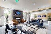 Located at 2550 Blackmon Dr in Decatur, GA, this single-floor amenity space offers a thoughtfully designed community lounge experience. The main lounge area serves as the centerpiece, anchored by a sleek wall-mounted fireplace framed in dark vertical paneling, flanked by cognac leather armchairs and a mounted flat-screen TV. Plush seating, including a navy blue sofa and bold patterned area rugs with geometric glass coffee tables, creates an inviting social atmosphere. Ambient cove lighting with warm golden accents along tray ceilings adds a modern, upscale feel throughout. Large floor-to-ceiling windows and glass double doors flood the space with natural light and provide views of the surrounding greenery. A dedicated games area features a contemporary stainless steel pool table set against a deep navy accent wall, also equipped with a wall-mounted TV and ambient pendant lighting. A built-in wet bar with stainless steel appliances and a sink offers convenient entertaining functionality. A high-top bar table with stools provides additional seating. The space also includes a private golf simulator room with a full projection screen and turf mat. Stylish pendant light fixtures, tall indoor plants, and mixed flooring of tile, carpet, and hardwood complete the polished, modern aesthetic of this versatile amenity space.