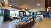 Located at 2 N Main St in Mount Prospect, Illinois, this single-floor amenity lounge is a thoughtfully designed social space that blends comfort and entertainment. The open-concept layout is anchored by a generously sized lounge area furnished with contemporary sofas and accent chairs arranged around glass coffee tables, complemented by curated artwork and lush indoor plants throughout. Large floor-to-ceiling windows line the perimeter, filling the space with natural light and offering pleasant views of the surrounding greenery. The herringbone-patterned hardwood flooring adds a refined architectural touch that flows seamlessly across the entire space. A full-size billiards table serves as a central focal point in the entertainment zone, while a stylish bar area features dramatic pendant lighting, bar-height seating, and a striking decorative back wall with a large display screen. A wood-paneled ceiling accent over the bar area introduces warmth and texture, contrasting beautifully with the deep navy-blue accent walls found throughout. A large-format television is mounted on a light stone feature wall in the lounge area, creating an ideal media viewing nook. Recessed and pendant lighting throughout ensures a warm, inviting ambiance suited for both casual gatherings and lively social events. Staircase access is also visible, connecting to additional areas of the building.