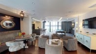 This single-floor lounge space is thoughtfully designed to blend elegance with functionality, offering multiple distinct zones for entertainment and relaxation. The open-concept layout flows seamlessly from one area to the next, anchored by rich hardwood flooring that grounds the overall design.

The lounge area features plush, neutral-toned barrel chairs and tufted seating arranged around low round tables, creating an inviting space for casual conversation. A decorative chandelier adds a refined touch near the dining area, which includes a circular table with upholstered chairs.

A sleek bar with dark, studded cabinetry and bar stool seating serves as a central focal point, ideal for entertaining. Adjacent to the bar, a billiards table sits within a carpeted game zone, framed by floor-to-ceiling windows that allow generous natural light to fill the space and offer views to the exterior.

Recessed lighting and elegant cove ceiling details with integrated LED strips add architectural interest overhead. Drapery in muted gray tones provides privacy when desired. The overall ambiance is contemporary and polished, suited for upscale communal use.