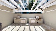 This outdoor amenity space is designed for communal entertaining and al fresco dining. The centerpiece of the area is a pair of built-in stainless steel grilling stations, each set into a sturdy concrete surround with cabinet storage below, providing ample prep and cooking space for residents and guests. The grilling stations are positioned side by side against a smooth stucco wall, offering an organized and functional outdoor kitchen setup.

Overhead, a striking white pergola with bold, radiating beams creates a bold geometric canopy that provides partial shade while maintaining an open, airy atmosphere. Two ceiling fans are mounted beneath the pergola, ensuring comfortable airflow during warm weather. The paved courtyard-style flooring features a classic brick-tone tile pattern, adding a refined texture to the outdoor setting.

Tall palm trees rise in the background, contributing a distinctly tropical ambiance to the space. The surrounding multi-story building facade, with its vibrant teal-accented balconies and large windows, frames the area and reinforces the resort-like character of the community. Natural light floods the space, casting dramatic linear shadows through the pergola beams, enhancing the visual appeal of this well-appointed outdoor gathering area.