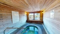 This single-level indoor spa room is a self-contained relaxation facility designed with a warm, lodge-inspired aesthetic. The space is fully enveloped in horizontal wood plank paneling, extending from floor to ceiling, creating a cohesive and inviting atmosphere throughout. A smooth wood-slat ceiling fitted with recessed lighting and wall-mounted accent lights provides a soft, warm glow that complements the natural tones of the room.

At the center of the space sits a built-in, oval-shaped hot tub with blue tile surround detailing and textured concrete decking. Stainless steel handrails are positioned at the entry point of the hot tub for safe and easy access. The overall layout is compact yet functional, allowing comfortable movement around the perimeter of the pool.

One wall features large, multi-pane windows with a built-in wooden bench seat beneath them, allowing an abundance of natural light to fill the room while offering views to the surrounding outdoor greenery and neighborhood. The windows contribute to an open, airy feel despite the enclosed nature of the space.

Posted signage includes a detailed list of spa rules and safety notices, indicating this is a shared amenity facility. The combination of natural wood materials, soft lighting, and a welcoming hot tub makes this spa room a functional and relaxing retreat.
