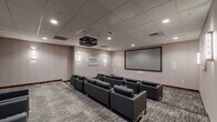 This single-floor screening and media lounge room offers a polished, purpose-built environment designed for comfortable group viewing and social gatherings. The room is finished with warm, neutral-toned walls and a suspended acoustic tile ceiling fitted with recessed lighting and a sleek, geometric ceiling-mounted projector that anchors the space visually. A large projection screen dominates one wall, providing an immersive viewing experience for all seated guests.

The seating is arranged in a theater-inspired configuration, featuring multiple rows of contemporary gray upholstered sofas and matching armchairs with clean, modern lines and chrome-finished legs. The seating is positioned to ensure unobstructed sightlines to the screen. Patterned, textured carpeting in neutral tones covers the floor throughout, adding warmth and sound absorption to the room.

Wall-mounted sconce lighting flanks framed artwork on the walls, contributing to a refined, ambient atmosphere. A small service area with cabinetry is visible toward the rear of the space, offering added functionality for events. The room is accessible via a solid wood door and includes clearly marked emergency exits, reflecting thoughtful attention to both design and safety. Overall, the space exudes a sophisticated yet inviting character ideal for screenings, presentations, or private events.