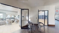 Located at Cortland Portofino in West Palm Beach, Florida, this expansive single-floor amenity facility offers a well-rounded mix of recreational, social, and functional spaces. The entry foyer features wood-look tile flooring and glass-paneled doors that provide clear sightlines into the adjoining areas, creating a welcoming and open first impression.

The centerpiece of the facility is a soaring full-size indoor basketball court with a polished hardwood floor marked with regulation court lines. Padded blue and gray wall panels line the perimeter for safety, while a row of clerestory windows along the upper walls floods the space with natural light. A mounted backboard and hoop complete the court, which also includes a built-in water fountain station along the sideline.

Adjacent to the court, a generously sized lounge area offers comfortable sectional seating, accent chairs, and a large wall-mounted TV set against a textured feature wall. Recessed lighting and light tile flooring contribute to the bright, modern aesthetic. A games nook with a foosball table and floor-to-ceiling windows overlooking lush palm-lined grounds adds a casual recreational element.

A compact workspace features a central stone-topped counter with bar seating, pendant lighting, dual wall-mounted screens, and garden views — well-suited for focused work or small gatherings. Throughout, the design maintains a cohesive contemporary palette of neutral tones accented with blues and warm wood details.