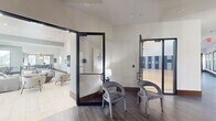 Located at Cortland Portofino in West Palm Beach, Florida, this expansive single-floor amenity facility offers a well-rounded mix of recreational, social, and functional spaces. The entry foyer features wood-look tile flooring and glass-paneled doors that provide clear sightlines into the adjoining areas, creating a welcoming and open first impression.

The centerpiece of the facility is a soaring full-size indoor basketball court with a polished hardwood floor marked with regulation court lines. Padded blue and gray wall panels line the perimeter for safety, while a row of clerestory windows along the upper walls floods the space with natural light. A mounted backboard and hoop complete the court, which also includes a built-in water fountain station along the sideline.

Adjacent to the court, a generously sized lounge area offers comfortable sectional seating, accent chairs, and a large wall-mounted TV set against a textured feature wall. Recessed lighting and light tile flooring contribute to the bright, modern aesthetic. A games nook with a foosball table and floor-to-ceiling windows overlooking lush palm-lined grounds adds a casual recreational element.

A compact workspace features a central stone-topped counter with bar seating, pendant lighting, dual wall-mounted screens, and garden views — well-suited for focused work or small gatherings. Throughout, the design maintains a cohesive contemporary palette of neutral tones accented with blues and warm wood details.