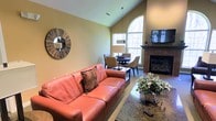 This single-floor residence offers a thoughtfully designed open-concept layout that seamlessly blends the living and dining areas into one cohesive, welcoming space. The living room is anchored by a bold coral leather sofa and accented with decorative touches, including a striking sunburst mirror that adds a artistic focal point to the warm, neutral-toned walls. A wall-mounted flat-screen television sits above a sleek, dark-mantled fireplace, providing both style and comfort to the room.

The soaring vaulted ceilings amplify the sense of openness and volume throughout the main living area, while a large arched window floods the space with abundant natural light, creating a bright and airy atmosphere. The dining area, positioned adjacent to the living room, features a round table with upholstered chairs and a contemporary pendant light fixture, making it an ideal spot for both casual meals and entertaining.

Granite-surface accents and polished floors contribute to the overall refined character of the space. Lush indoor greenery adds a natural, refreshing element that complements the warm interior palette. Together, these features create a space that is both functional and inviting, offering a comfortable and visually appealing environment throughout.