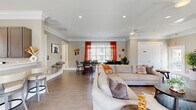 Located at 831 Pecan Ave in Winston-Salem, NC, this single-floor community space offers a well-designed, open-concept layout that seamlessly connects the living, dining, and kitchen areas. The living room is anchored by a large sectional sofa and features recessed lighting, crown molding, a ceiling fan, and durable tile flooring throughout. A wall-mounted flat-screen TV and a shuffleboard table define a dedicated game and lounge area, complemented by a glass-paned exterior door that brings in natural light and outdoor views. The dining area accommodates a large, dark-wood trestle table with ample seating, bathed in natural light from wide windows dressed with bold orange curtains. The kitchen is outfitted with gray shaker-style cabinetry, light quartz countertops, stainless steel appliances including a range, built-in microwave, and dishwasher, plus a deep undermount sink beneath a bright window. Two bathrooms are finished with tile flooring and include accessibility features such as grab bars. Crown molding, neutral warm-gray walls, and consistent tile flooring unify the space, creating a clean, welcoming atmosphere suited for communal gatherings and everyday use.
