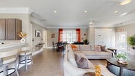 Located at 831 Pecan Ave in Winston-Salem, NC, this single-floor community space offers a well-designed, open-concept layout that seamlessly connects the living, dining, and kitchen areas. The living room is anchored by a large sectional sofa and features recessed lighting, crown molding, a ceiling fan, and durable tile flooring throughout. A wall-mounted flat-screen TV and a shuffleboard table define a dedicated game and lounge area, complemented by a glass-paned exterior door that brings in natural light and outdoor views. The dining area accommodates a large, dark-wood trestle table with ample seating, bathed in natural light from wide windows dressed with bold orange curtains. The kitchen is outfitted with gray shaker-style cabinetry, light quartz countertops, stainless steel appliances including a range, built-in microwave, and dishwasher, plus a deep undermount sink beneath a bright window. Two bathrooms are finished with tile flooring and include accessibility features such as grab bars. Crown molding, neutral warm-gray walls, and consistent tile flooring unify the space, creating a clean, welcoming atmosphere suited for communal gatherings and everyday use.