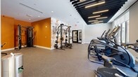 Located at 100 Hamilton St in Bound Brook, NJ, this fitness center offers a thoughtfully designed exercise space spread across a single floor. The facility is divided into distinct workout zones, each catering to different training needs. The cardio area features a row of treadmills and elliptical machines positioned along large windows that flood the space with natural light and provide views of the surrounding streetscape. A striking slatted black ceiling with integrated linear LED lighting runs throughout, lending the space a modern, industrial aesthetic. The strength training zone is anchored by a comprehensive dumbbell rack, adjustable benches, and cable machines, complemented by full-length mirrors that visually expand the area. Bold orange accent walls and a motivational graphic mural — reading "The Stronger You Are, The Better You Feel" — add energy and character to the environment. A separate, glass-partitioned room provides additional space for functional training or dedicated machine work. Rubber flooring throughout ensures durability and safety across all workout areas. The overall design balances high-end finishes with practical functionality, creating a professional-grade fitness environment that is both visually dynamic and comprehensively equipped for a wide range of exercise routines.