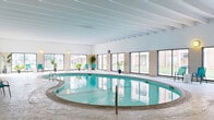Located at 100 Thompson Ave W in West St. Paul, Minnesota, this single-floor community amenity facility offers a generously sized, well-appointed space designed for both recreation and gatherings. The centerpiece of the facility is a large, freeform indoor swimming pool with striking turquoise water, set beneath an architecturally distinctive ceiling featuring exposed white structural beams. An expansive wall of windows and sliding glass doors surrounds the pool area, flooding the space with natural light and providing open views of the surrounding green grounds. Seating areas with teal accent chairs and tropical potted plants add a welcoming, resort-like atmosphere to the poolside. Adjacent to the pool is a bright sunroom-style lounge area, also benefiting from floor-to-ceiling windows and outdoor views, offering a comfortable space to relax. The facility also includes a carpeted multi-purpose community room equipped with long tables and chairs, suitable for meetings or events, along with a secondary room featuring a small kitchenette area with a refrigerator. A hallway connects the various areas, completing the functional layout. Together, these spaces create a versatile, amenity-rich environment suited for recreation and community use.