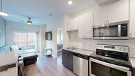 Located at 300 W State St in West Lafayette, Indiana, this 4-bedroom, 3-bathroom unit offers a functional and modern layout across a single floor. The kitchen is outfitted with stainless steel appliances — including a refrigerator, range, microwave, and dishwasher — complemented by white upper cabinetry, dark lower cabinets, a subway tile backsplash, and light countertops. Wood-look flooring runs throughout the main living areas, adding warmth and cohesion to the space. The combined kitchen and living area flows into a comfortable living room featuring a sliding glass door that opens to a private balcony with views of the surrounding streetscape. Each bedroom is designed with practicality in mind, featuring windows with blinds that allow natural light, ceiling fans, and access to closets with built-in shelving and dresser storage. The bathrooms are finished with penny-tile flooring, subway tile surrounds, dark wood vanities, and combination tub/shower units, offering a clean and contemporary aesthetic. The neutral color palette throughout the unit — soft whites, warm beiges, and light grays — creates a bright and cohesive atmosphere well-suited for shared living.