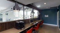 This single-floor business center offers a functional and well-appointed workspace designed for productivity. A long, continuous desk runs along a wall of large windows, providing abundant natural light and views of the adjacent fitness area, which is visible through the glass partition. The workstation area is equipped with desktop computers and a printer, making it suitable for a range of professional tasks. Vibrant red ergonomic chairs add a pop of color against the dark wood paneling and deep teal accent wall, creating a modern, energetic aesthetic. Overhead, a series of industrial-style pendant lights hang at varying heights, complementing the contemporary design while ensuring ample task lighting throughout the space. The neutral carpeted flooring grounds the room, balancing the bold color choices. The open sightlines into the fitness facility beyond reinforce the communal nature of the space, suggesting it is part of a larger amenity-rich building. Overall, the business center combines a clean, modern design with practical features, offering an inviting environment for focused work.