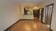 This single-floor unit is designed with an open-concept layout that connects the living area to the kitchen seamlessly. Warm-toned hardwood flooring extends throughout the main living space, providing a cohesive and inviting aesthetic. The kitchen features dark espresso cabinetry, a stainless steel refrigerator, and a peninsula with a contrasting light-colored base, offering additional counter space and a natural division between the kitchen and living areas. A modern pendant light fixture with multiple hanging lights is positioned above the kitchen peninsula, serving as a functional and decorative focal point. The walls are finished in a neutral tone, complementing the dark trim and baseboards that frame the space. A hallway visible from the main area leads to additional rooms, with dark-stained doors maintaining design consistency throughout. Notably, in-unit laundry is included, with a washer and dryer visible behind a slatted door near the hallway. The layout is practical and efficient, with clearly defined zones for living, cooking, and utility needs, making it a well-organized single-floor space.