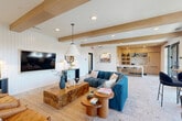 Located at 110 E 36th Street in Charlotte, NC, this single-floor lounge space offers a thoughtfully designed living area that blends coastal charm with modern sophistication. The room is anchored by striking exposed natural wood ceiling beams that contrast beautifully against crisp white surfaces, including distinctive shiplap accent walls. Recessed lighting and a statement pendant light contribute to a warm, inviting ambiance throughout.

The seating area is furnished with a mix of tufted caramel leather chairs and a deep teal sectional sofa, centered around a substantial live-edge wood block coffee table that serves as a focal point of the room. Complementary round wooden side tables and curated coastal-inspired décor, including ceramic vases and framed artwork, add layers of visual interest.

Large floor-to-ceiling windows with black steel frames line one side of the space, flooding the interior with abundant natural light and providing direct access to an outdoor terrace. A built-in wet bar area features light wood cabinetry, marble countertops, and teal pendant lighting, adding a functional and elegant touch. Stone tile flooring flows seamlessly throughout the open layout. Curated art pieces, including a large flamingo photograph and architectural prints, complete the refined yet relaxed aesthetic of this well-appointed lounge.