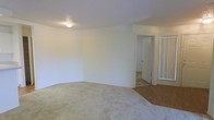 This single-floor space presents a clean, minimalist layout with a neutral design palette. The main living area is finished with wall-to-wall carpet in a light beige tone, providing a soft underfoot feel and a warm, understated aesthetic. The white walls and ceiling create a bright, open atmosphere, enhanced by a centrally mounted ceiling light fixture with a textured ceiling finish visible overhead.

A notable flooring transition from carpet to hard surface flooring is visible toward the rear of the space, delineating a separate functional zone — likely a kitchen, entryway, or utility area. A doorway leads to an additional room beyond, suggesting the layout extends further and offers dedicated spaces for various uses.

A mirrored sliding closet door is partially visible along one wall, indicating practical built-in storage integrated into the design. The open doorway and interconnected layout contribute to a functional flow between spaces. Overall, the space offers a straightforward, move-in-ready interior with clean lines, ample wall space, and a simple yet functional design suited to a variety of furnishing arrangements.
