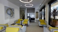 This contemporary single-floor lounge is designed with both comfort and functionality in mind. The space is anchored by a stylish seating area furnished with distinctive gray upholstered chairs accented by vibrant yellow cushions, arranged around small dark side tables to create intimate conversation groupings. Overhead, striking circular ring pendant lights add a modern focal point, complemented by recessed ceiling lighting that ensures the space is well-illuminated throughout.

A built-in kitchenette is neatly integrated into the background, featuring dark cabinetry, upper storage cabinets, a sink, and what appears to be a coffee or beverage station — providing convenient refreshment access for occupants.

Transitioning further into the space, a secondary seating area opens up with expansive floor-to-ceiling windows that invite generous natural light and offer views of lush outdoor plantings. The continuation of the circular pendant lighting creates a cohesive design language throughout. The flooring transitions from light-toned tile in the entry corridor to a neutral carpet in the lounge areas, subtly defining each zone. A staircase is visible at the rear of the main lounge, suggesting access to additional building areas. Overall, the space presents a clean, professional, and welcoming ambiance suited for community or hospitality use.