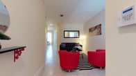 This two-story space offers a clean, contemporary layout designed for comfortable living. The main living area immediately draws attention with its light-toned tile flooring that runs throughout, creating a cohesive and airy feel. The room is furnished with a striking combination of a dark tufted sofa paired with vibrant red accent chairs, all anchored by a bold black-and-white patterned area rug — delivering a lively contrast against the neutral white walls. A wall-mounted mirror and curated artwork add personality and depth to the space. A floor lamp provides warm, ambient lighting that complements the natural light filtering through the windows. A long corridor extends toward additional rooms, hinting at a generous floor plan with multiple functional areas, including what appears to be a dining space visible in the background. The high, smooth ceilings enhance the sense of openness throughout. With its two floors, the property offers ample room across multiple levels, making it well-suited for a variety of living arrangements. The overall ambiance balances modern aesthetics with practical design, creating a welcoming and visually dynamic environment.