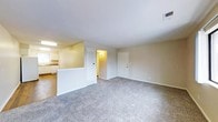 This single-floor residence features a practical and straightforward layout that connects the main living area to the kitchen through a half-wall partition, creating an open yet defined sense of separation between the two spaces. The living area is carpeted in a neutral gray tone, providing a soft, understated foundation that complements the light beige walls and white trim throughout. The smooth white ceiling enhances the brightness of the room, contributing to an airy feel.

A louvered closet door adjacent to the kitchen partition adds a classic touch and provides convenient storage within the living space. The kitchen, visible beyond the half-wall, features white cabinetry and appears well-lit with overhead fluorescent lighting, offering a functional workspace. A hallway leads toward additional rooms at the rear of the unit, illuminated by a wall-mounted light fixture, adding warmth to the interior corridor. The main entry door is positioned along the far wall of the living area, with a visible HVAC vent overhead ensuring climate control. Overall, the space presents a clean, move-in ready environment with a simple, cohesive aesthetic.
