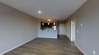 This single-floor unit showcases an open-concept layout that seamlessly connects the living area and kitchen, creating a spacious and functional environment. The kitchen is a standout feature, finished with rich dark cabinetry, light-toned countertops, and stainless steel appliances. A raised breakfast bar peninsula divides the kitchen from the living space, offering casual seating and additional prep area. Three pendant lights hang above the bar, providing focused task lighting while contributing to the modern aesthetic. Track lighting in the kitchen further enhances visibility throughout the cooking area. The flooring throughout the main space is a warm, medium-toned hardwood-style plank, offering durability and visual continuity. Walls are finished in a neutral warm grey tone, complementing the overall palette. Recessed lighting in the ceiling provides clean, ambient illumination. A textured ceiling adds subtle character to the space. A hallway visible from the living area leads to additional rooms, including what appears to be a closet and bathroom, maintaining a practical and well-organized floor plan. White trim and baseboards provide a crisp contrast throughout.