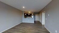 This single-floor unit showcases an open-concept layout that seamlessly connects the living area and kitchen, creating a spacious and functional environment. The kitchen is a standout feature, finished with rich dark cabinetry, light-toned countertops, and stainless steel appliances. A raised breakfast bar peninsula divides the kitchen from the living space, offering casual seating and additional prep area. Three pendant lights hang above the bar, providing focused task lighting while contributing to the modern aesthetic. Track lighting in the kitchen further enhances visibility throughout the cooking area. The flooring throughout the main space is a warm, medium-toned hardwood-style plank, offering durability and visual continuity. Walls are finished in a neutral warm grey tone, complementing the overall palette. Recessed lighting in the ceiling provides clean, ambient illumination. A textured ceiling adds subtle character to the space. A hallway visible from the living area leads to additional rooms, including what appears to be a closet and bathroom, maintaining a practical and well-organized floor plan. White trim and baseboards provide a crisp contrast throughout.