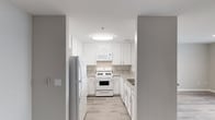 This single-floor space offers a clean, modern aesthetic highlighted by a well-appointed kitchen. The kitchen is designed with crisp white shaker-style cabinetry that extends to the ceiling, providing ample storage space. Light-toned granite-style countertops complement the white finish, while a classic subway-style backsplash ties the design together cohesively. The kitchen is equipped with a full suite of white appliances, including a gas range, over-the-range microwave, refrigerator, and a sink positioned along the side counter. Light wood-look flooring runs throughout, adding warmth and continuity to the space. A flush-mount ceiling light fixture ensures the kitchen is well-lit, contributing to the overall bright and airy atmosphere. The layout is efficiently organized, making practical use of the available space with a functional flow between the cooking, prep, and storage areas. Neutral gray tones on the surrounding walls provide a subtle contrast to the white cabinetry, creating a balanced and inviting interior palette. The overall design reflects a contemporary, move-in-ready aesthetic suited for everyday living.