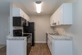 This single-floor residence in Greensboro, NC (27455) offers a well-organized layout with a thoughtful mix of flooring and finishes throughout. The living room serves as a central gathering space, featuring plush carpeting, a wood-burning fireplace, and a sliding glass door framed by vertical blinds that allows natural light to fill the room. The transition between the living area and entry is marked by warm wood-look flooring, which also extends into the kitchen and hallways. The galley-style kitchen is efficiently designed with crisp white shaker cabinets, granite-inspired countertops, a double sink, and black appliances including a range, microwave, and refrigerator. The bedrooms are carpeted and finished with neutral tones, offering a comfortable and quiet atmosphere. At least one bedroom features a ceiling fan with light, a reach-in closet, and direct access to an adjacent room. The bathroom is outfitted with wood-look flooring, a white vanity, a towel ring, and a grab bar for added accessibility. Textured ceilings are consistent throughout the unit, contributing to its classic residential character. Overall, this space presents a clean, functional, and move-in-ready environment.