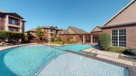 This residential community showcases classic brick architecture with pitched rooflines, multi-story buildings, and a warm, traditional aesthetic throughout. The exterior is defined by rich red and tan brick facades, accented by large windows, private balconies, and carefully maintained landscaping that includes mature trees, sculpted shrubs, and lush green lawns.

At the heart of the community lies a spacious resort-style swimming pool with crystal-clear blue water, complemented by a generously sized brick-paved pool deck. The deck is furnished with lounge chairs ideal for sunbathing, along with shaded patio seating areas fitted with umbrellas and tables, creating an inviting space for both relaxation and socializing. A pergola structure adds architectural interest and additional shade near the pool area.

Winding pedestrian pathways connect the various buildings and amenity areas, framed by vibrant seasonal plantings and well-groomed garden beds. The grounds maintain a cohesive, polished appearance throughout. Multiple outdoor seating vignettes are positioned around the community, offering residents comfortable spaces to gather or unwind. The overall ambiance of the outdoor spaces is that of a serene, resort-inspired retreat, combining functional amenities with thoughtfully designed landscaping to create an attractive and livable community environment.