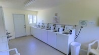 This single-floor communal laundry facility is designed for straightforward, efficient use. The room is laid out with a long row of top-loading washing machines lined against one wall, providing ample capacity for multiple users. The machines are uniformly white and neatly arranged, reflecting a well-kept and organized environment. A posted sign from management reminds users to keep the laundry facilities clean, indicating attentive property oversight.

The space features light-colored walls and a smooth floor surface that is easy to maintain and keep tidy. A window on the far wall allows natural light to filter into the room, complementing the overhead fluorescent lighting to keep the area bright and well-illuminated. A dedicated entry door provides direct access to the facility, ensuring convenience for users.

The overall ambiance is functional and utilitarian, prioritizing practicality and cleanliness. The open floor space in front of the machines allows for comfortable movement while loading and unloading laundry. A waste bin is positioned near the machines for easy disposal of detergent packaging and other waste, further supporting the cleanliness of the space. This laundry facility is a well-organized, accessible shared amenity suited to serving a residential community.