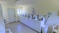 This single-floor communal laundry facility is designed for straightforward, efficient use. The room is laid out with a long row of top-loading washing machines lined against one wall, providing ample capacity for multiple users. The machines are uniformly white and neatly arranged, reflecting a well-kept and organized environment. A posted sign from management reminds users to keep the laundry facilities clean, indicating attentive property oversight.

The space features light-colored walls and a smooth floor surface that is easy to maintain and keep tidy. A window on the far wall allows natural light to filter into the room, complementing the overhead fluorescent lighting to keep the area bright and well-illuminated. A dedicated entry door provides direct access to the facility, ensuring convenience for users.

The overall ambiance is functional and utilitarian, prioritizing practicality and cleanliness. The open floor space in front of the machines allows for comfortable movement while loading and unloading laundry. A waste bin is positioned near the machines for easy disposal of detergent packaging and other waste, further supporting the cleanliness of the space. This laundry facility is a well-organized, accessible shared amenity suited to serving a residential community.