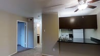 This single-floor unit showcases a practical, open-concept layout that connects the kitchen and living areas seamlessly. The kitchen is equipped with dark espresso-toned upper cabinetry, a contrasting dark granite-style countertop breakfast bar, a white electric range, and a white refrigerator. An overhead microwave or range hood is integrated above the stove, maximizing functionality in the cooking space. The kitchen's peninsula-style counter creates a natural division between the cooking area and the living space, while still maintaining an open and connected feel. The living area features wall-to-wall carpeting and is topped with a ceiling fan, providing both comfort and air circulation. A textured popcorn ceiling runs throughout, adding a classic residential character. Neutral beige and tan wall tones create a warm, cohesive ambiance across the space. A hallway extends from the main living area toward what appears to be a bathroom, visible in the background with its own lighting. The unit also includes visible HVAC vents, indicating a central air system. Overall, the layout is efficient and functional, offering a comfortable everyday living environment.