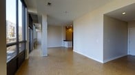 This single-floor space presents an open-plan layout with a clean, minimalist aesthetic. The main living area is generously proportioned, featuring polished aggregate flooring that adds texture and visual interest underfoot. Neutral white walls and white baseboards contribute to a bright, airy atmosphere throughout. A prominent structural column stands within the open area, serving as an architectural detail that subtly defines zones within the space. Recessed lighting is installed in the ceiling, providing even illumination across the interior. Toward the rear, a kitchen counter or bar area is partially visible, suggesting a semi-open kitchen configuration with warm-toned cabinetry and dark countertops. On the left side of the space, large floor-to-ceiling windows or glazed panels allow natural light to filter in, enhancing the sense of openness. Ceiling-mounted HVAC vents are integrated throughout, ensuring climate control functionality. The overall ambiance is understated and versatile, with an unfinished quality that suggests the space is ready for customization or fit-out to suit a variety of uses.