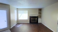 This single-floor space presents a clean, functional layout with a welcoming living area as its centerpiece. The living room is anchored by a classic fireplace finished with dark tile surround and a white mantel, adding a focal point of warmth and character to the room. Flanking the fireplace are built-in shelves and a small cabinet, providing convenient storage and display options. Rich hardwood flooring extends throughout, lending a sense of warmth and continuity to the space. A large window along the adjacent wall allows ample natural light to fill the room, brightening the neutral, beige-toned walls. The textured popcorn ceiling adds a traditional touch to the overall aesthetic. The open, uncluttered layout offers flexibility for a variety of furniture arrangements, making it easy to personalize the space. White baseboards and trim provide clean contrast against the darker flooring, contributing to a cohesive interior finish. Overall, the space combines practical features with comfortable, everyday livability in a straightforward single-level setting.
