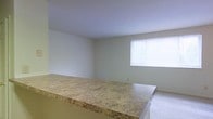 This single-floor space features an open-concept layout that connects the kitchen and living areas seamlessly. A prominent breakfast bar with a granite-patterned laminate countertop serves as the dividing element between the two zones, offering both a casual dining surface and a functional workspace. The countertop's warm beige and gold tones add a subtle visual anchor to the otherwise neutral interior.

The living area is finished with light-colored carpeting, contributing to a clean and understated aesthetic. The walls are painted in a soft, neutral tone throughout, creating a cohesive and versatile backdrop suitable for a variety of furnishing styles. A window on the far wall allows natural light to filter into the living space, brightening the room and providing ventilation. The window is fitted with horizontal blinds, offering adjustable light control and privacy.

The ceilings are smooth and painted white, maintaining a simple, uncluttered appearance. Overall, the space presents a functional and practical layout with clean finishes, making it well-suited for everyday living.